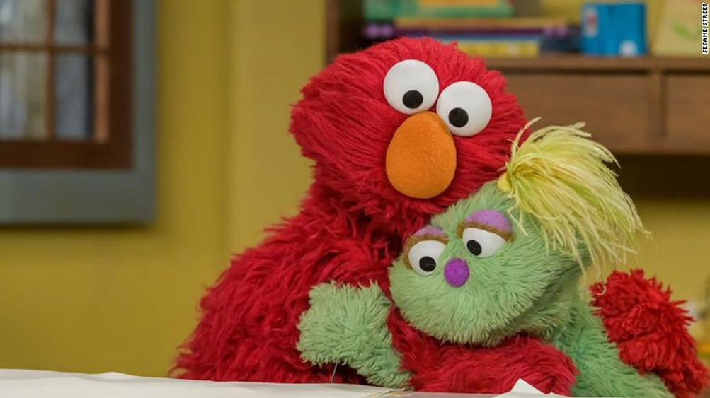 Elmo hugging a green puppet friend with yellow hair, both sitting together in a warm indoor setting.