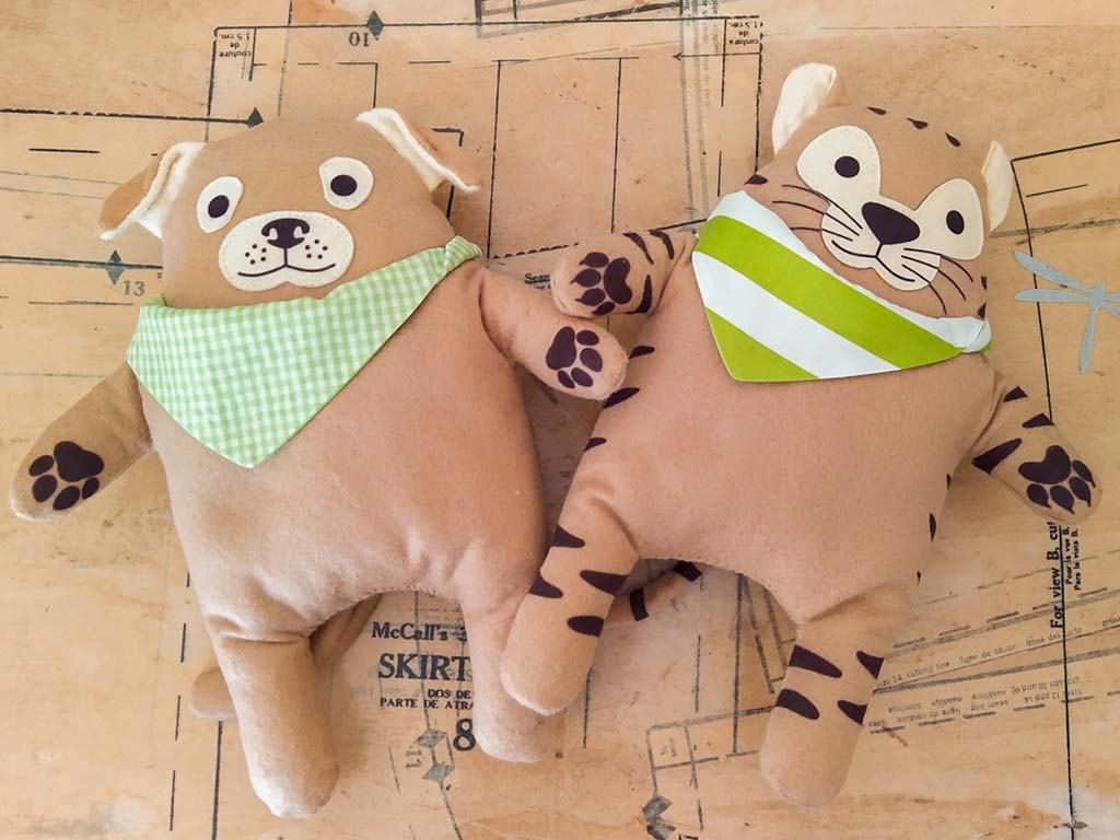 Two handmade plush toys—a dog and a cat—wearing green patterned bandanas lying on a sewing pattern table.