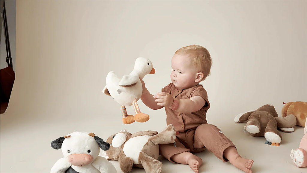 A baby sitting on the floor holding a white plush goose while surrounded by several soft stuffed animal toys.