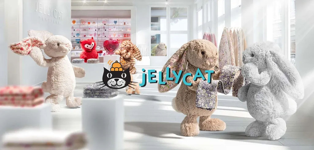 Jellycat bunnies and plush animals posed inside a bright, modern store, holding soft blankets and fabrics, with the Jellycat logo displayed in the center and shelves of colorful plush toys in the background.