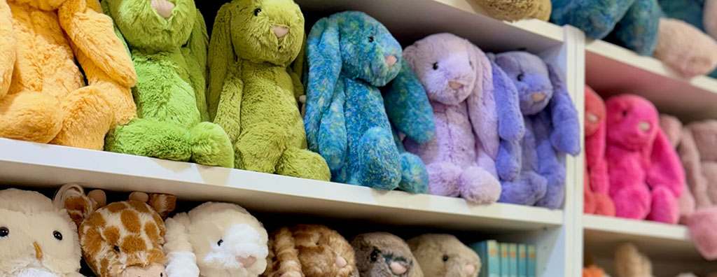 Shelves filled with plush toys arranged by color, including green, blue, purple, and pink bunnies, along with various animal plushies on the lower shelf in a cozy store setting.