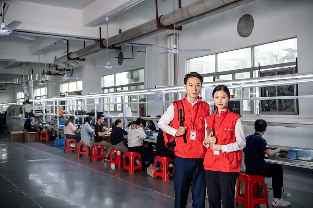 Two quality inspectors wearing red vests stand inside a plush toy manufacturing workshop, with workers on production lines assembling products in a clean, organized factory environment.