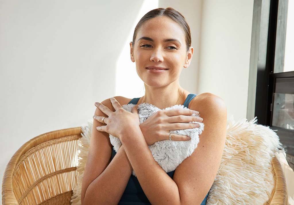 A woman sitting in a wicker chair smiling softly while hugging a fluffy gray plush heating pad to her chest in a cozy, sunlit room.