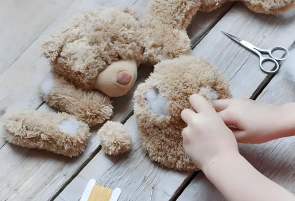 Hands repairing a torn teddy bear, restuffing the plush toy with fiberfill on a wooden table.
