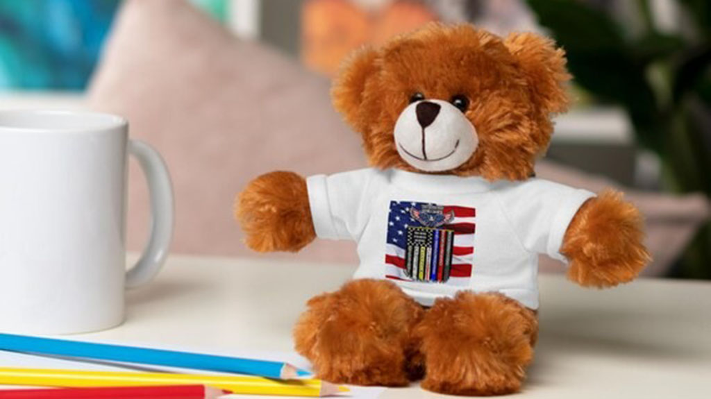 Soft brown teddy bear wearing a white T-shirt printed with an American flag design, sitting on a table beside a coffee mug and colored pencils.