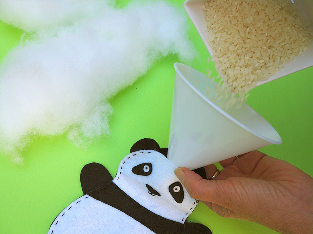 Close-up of hands using a funnel to pour rice into a handmade felt panda plush, with polyester stuffing in the background on a bright green surface.