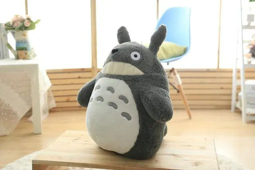 Grey Totoro plush toy sitting on a wooden table in a bright cozy room with soft natural lighting.