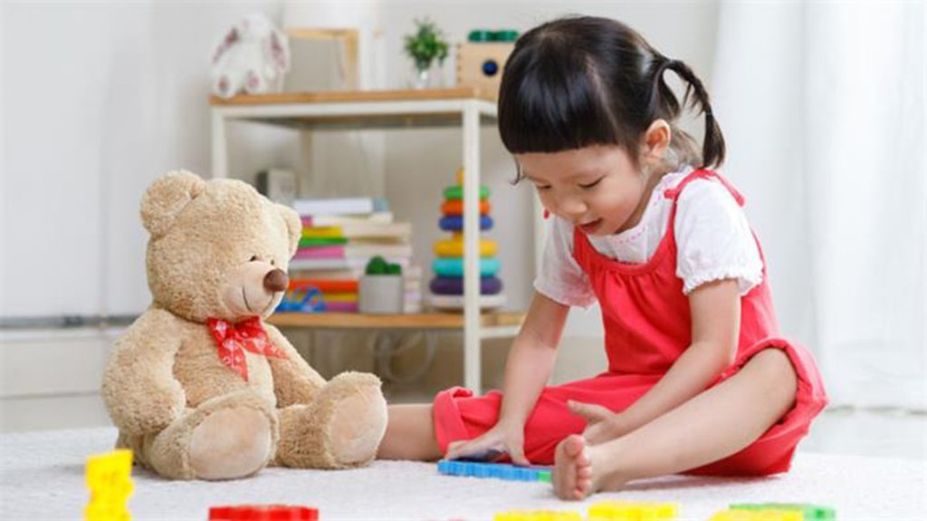 Young girl sitting on the floor playing with colorful blocks beside a plush teddy bear wearing a red ribbon.