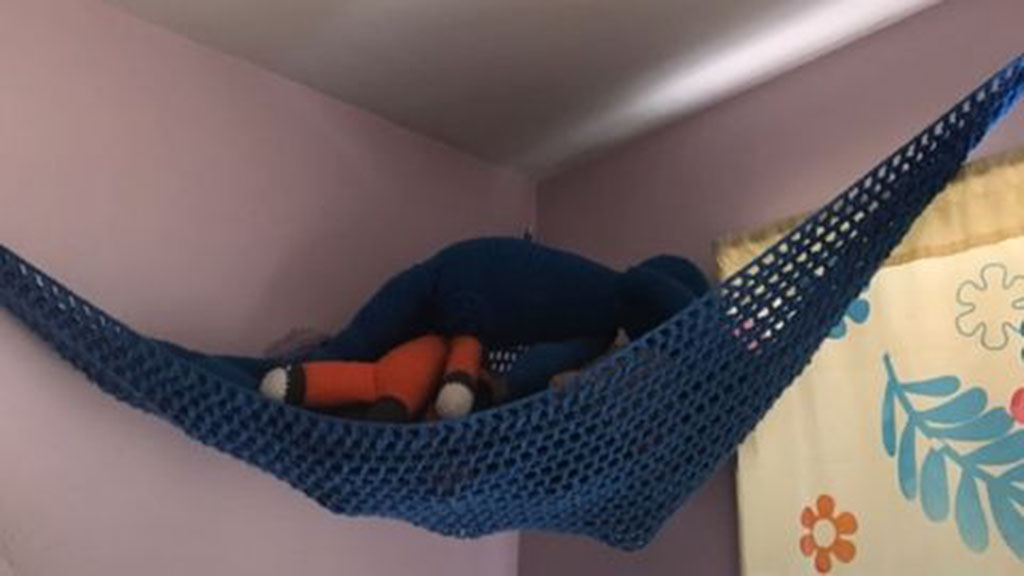 Blue crocheted stuffed animal hammock hanging in a room corner holding plush toys