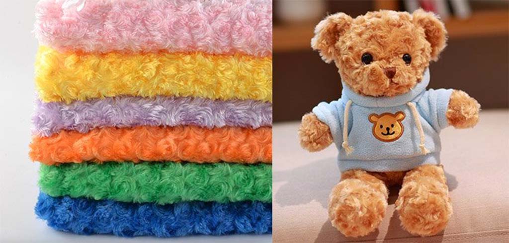 Stack of colorful rose fleece plush fabrics beside a soft teddy bear wearing a blue hoodie, showing material used for custom stuffed toys.
