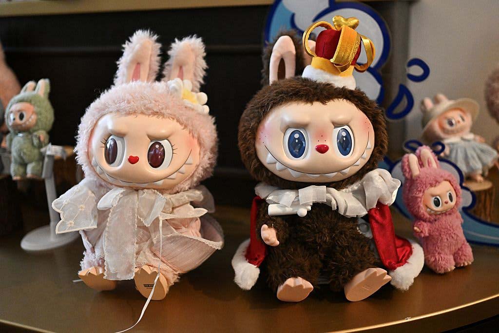 Two Labubu-style plush dolls displayed side by side, one dressed in a pink lace outfit with bunny ears and the other in a brown royal-themed costume with a crown. Smaller matching plush figures are arranged in the background on a decorative shelf.