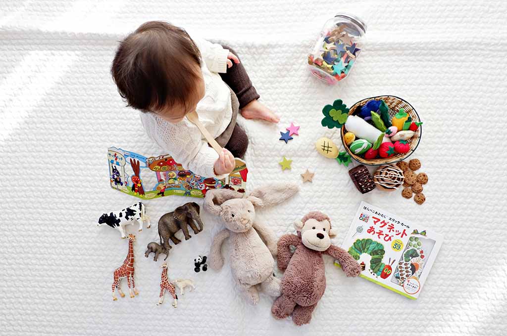 Baby sitting on a white blanket surrounded by plush toys, animal figurines, fabric food toys, wooden stars, and picture books, engaging in sensory and imaginative play.