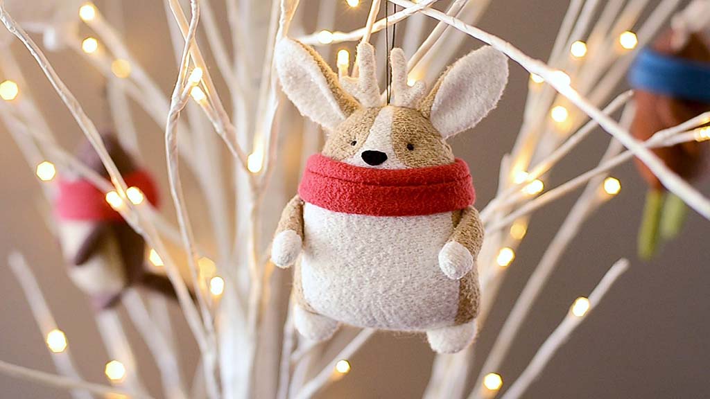 Soft corgi plush ornament wearing a red scarf, hanging on a white decorative branch with warm fairy lights in the background, styled for holiday or winter decoration.