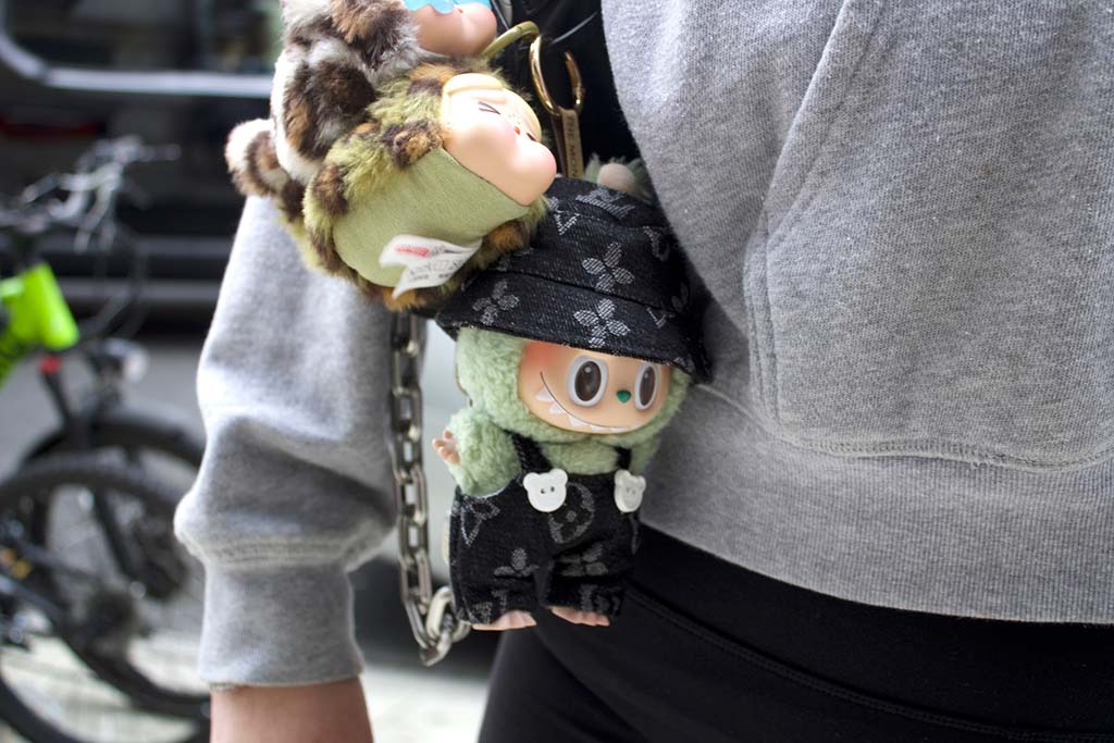 Close-up of a person wearing a gray sweatshirt and black pants with two Labubu plush keychains attached to a chain strap bag, featuring designer-inspired outfits.