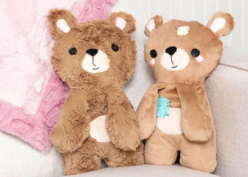 Two handmade plush bear toys with stitched faces and button eyes sitting on a sofa, one fuzzy and one smooth, beside a pink blanket.