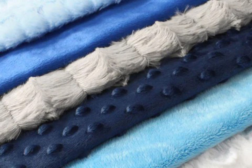Close-up view of assorted minky plush fabrics in blue and gray tones featuring smooth, dotted, and long-pile textures.