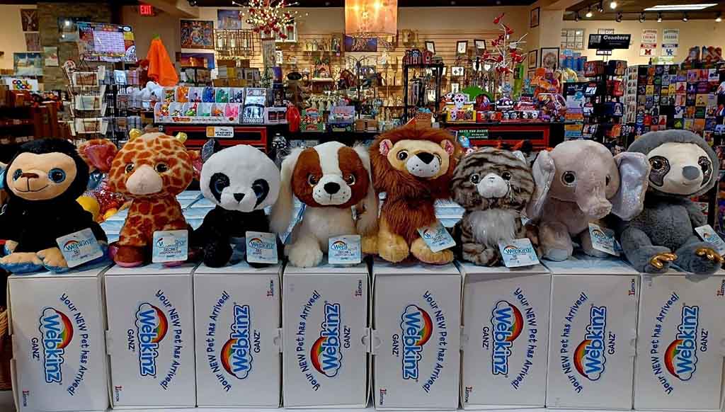 A row of Webkinz plush toys—including a monkey, giraffe, panda, dog, lion, cat, elephant, and sloth—lined up on top of Webkinz boxes inside a toy store.