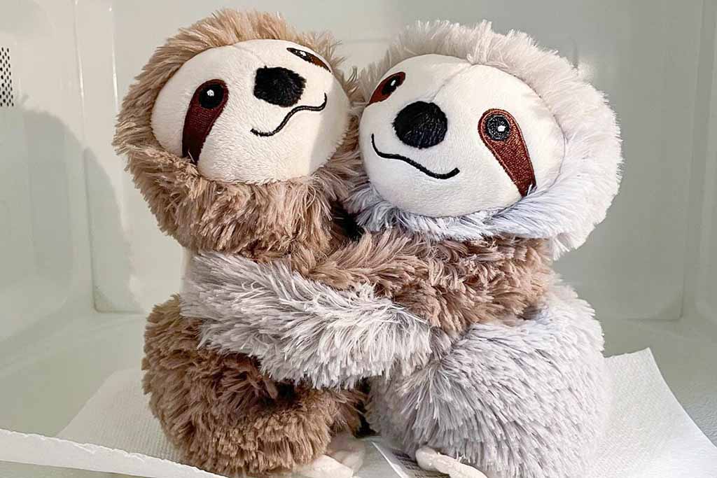 Two fluffy sloth plush toys hugging each other, one brown and one light gray, sitting upright on a white surface.