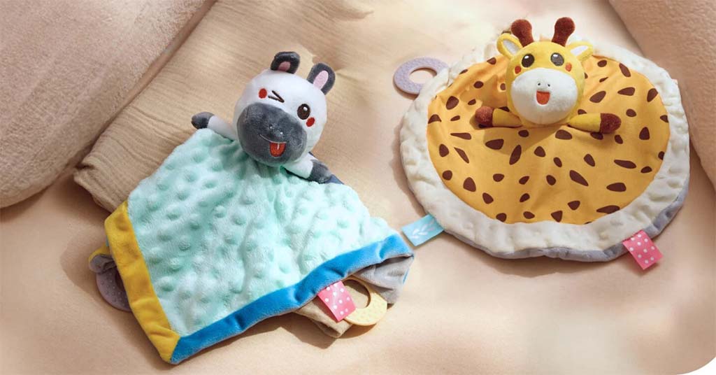 Soft baby comforter toys featuring a hippo and a giraffe plush design, each attached to a cuddly blanket with colorful tags, displayed on a beige bedding background.