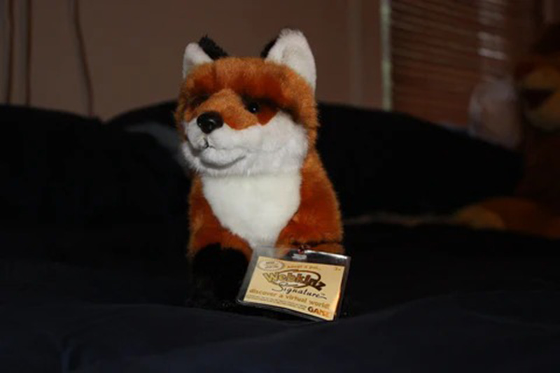 A Webkinz Signature fox plush toy sitting on a dark bedspread with its tag displayed in front.