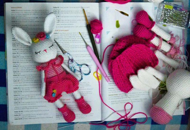 Handmade amigurumi bunny parts, crochet tools, yarn, and pins laid out on an open pattern book during plush toy assembly.