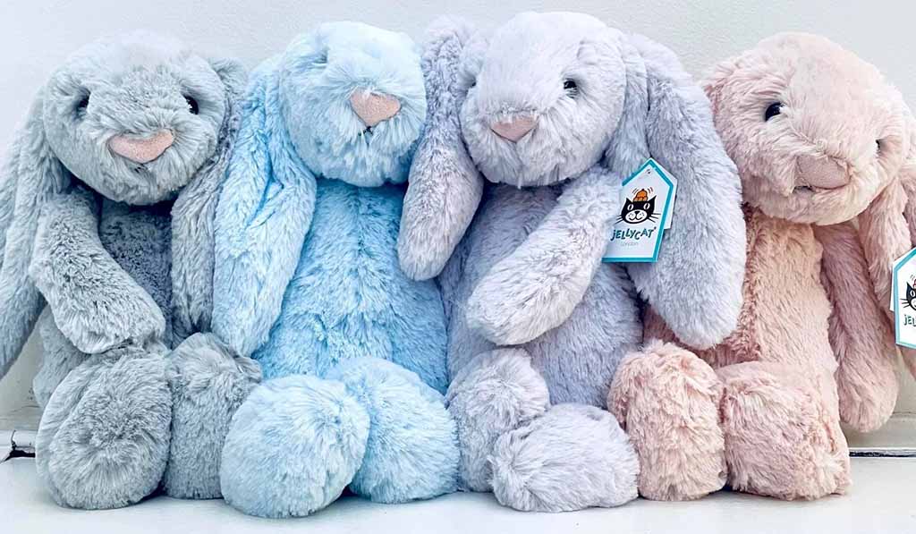A row of four soft pastel-colored plush bunny toys sitting side by side, featuring long floppy ears, embroidered faces, and Jellycat-style tags, displayed against a light background.