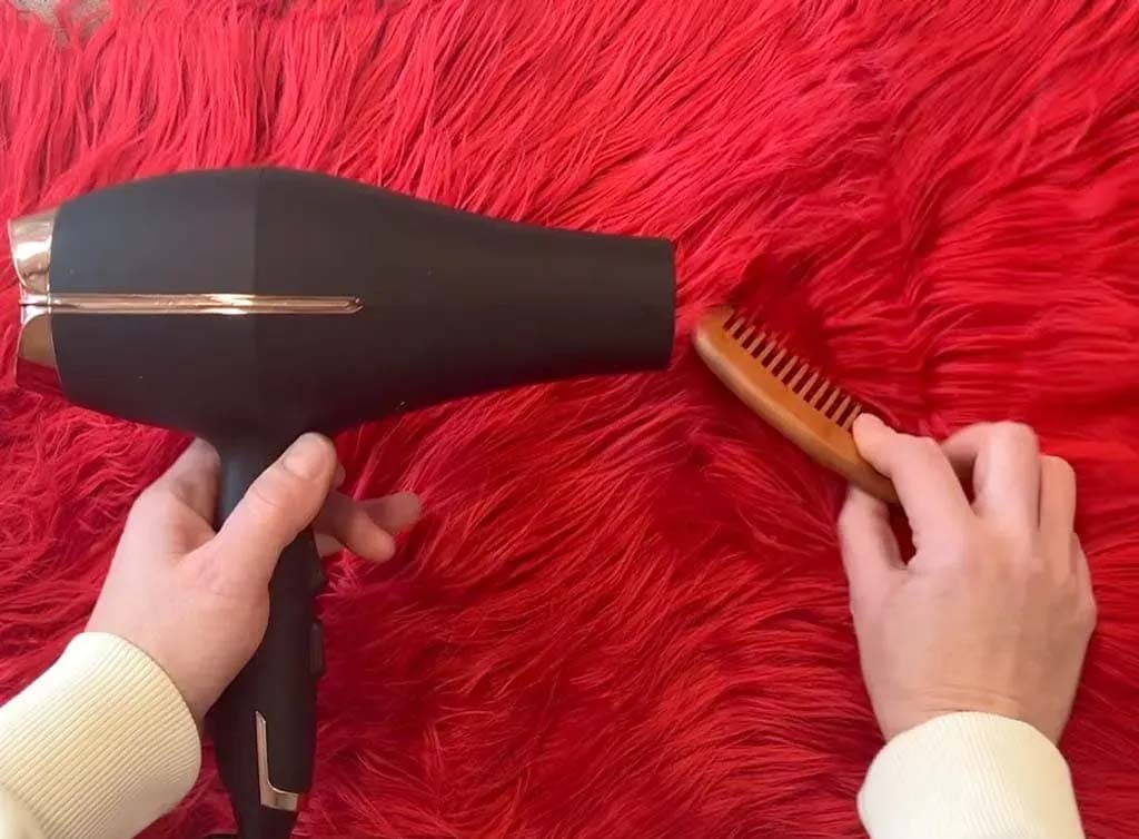 Hands using a hairdryer and wooden comb to smooth red faux fur fabric, demonstrating proper care and fluffing technique for synthetic fur.
