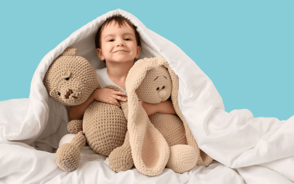 Smiling child sitting under a white blanket, hugging two crocheted plush toys — a bear and a bunny — against a light blue background.