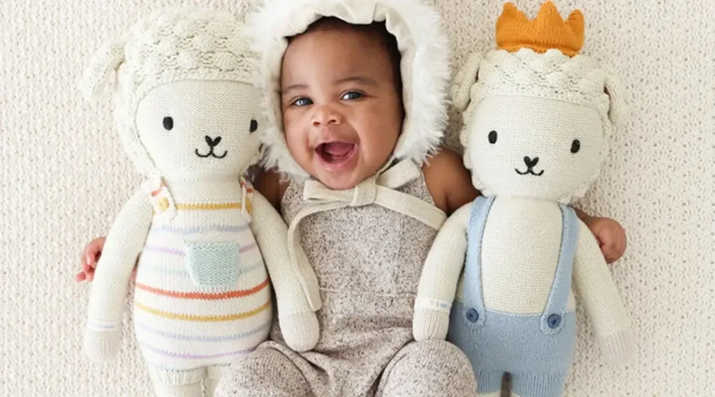 Happy baby lying between two soft knitted plush dolls, smiling brightly on a cozy textured blanket.