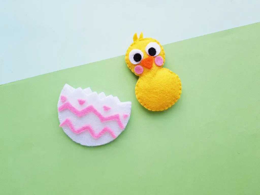 Handmade felt craft pieces including a yellow chick with large eyes and a pink-decorated broken egg shell placed on a pastel green background.