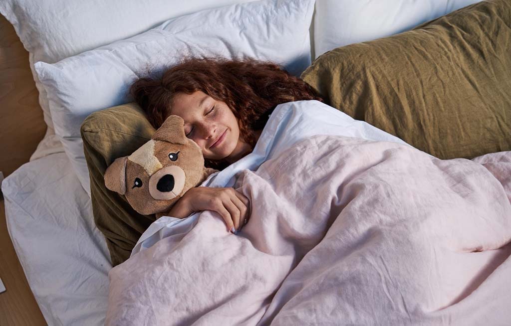A woman with curly hair sleeps peacefully in bed, cuddling a soft brown and beige plush dog under a pink blanket.