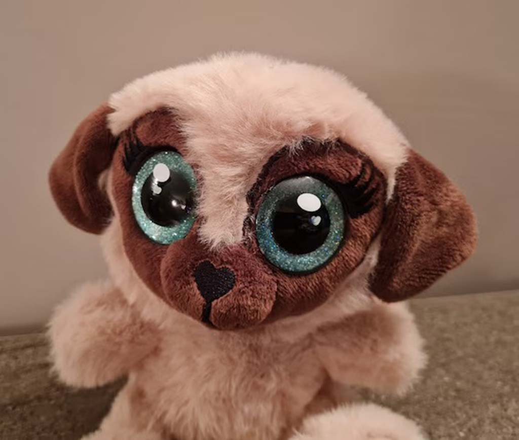 A soft beige pug plush toy with large glittery turquoise eyes, brown ears, and a heart-shaped nose, sitting upright against a neutral background.
