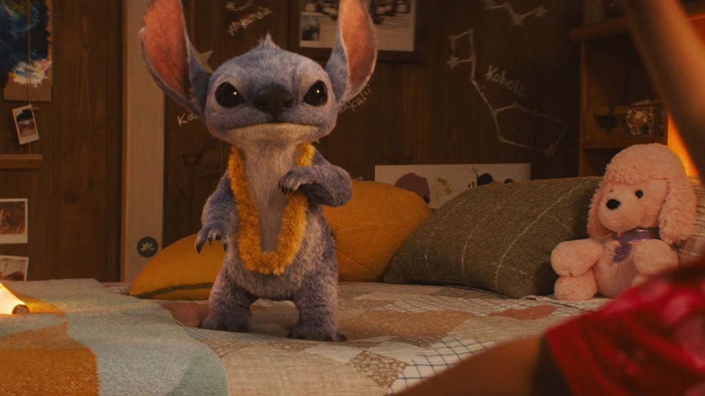 A small blue Stitch-like animated character wearing a yellow lei stands on a cozy bedroom bed decorated with pillows, plush toys, and warm lighting.