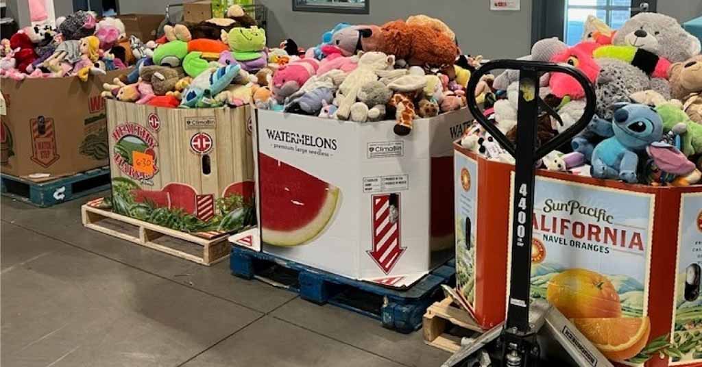 Large pallet bins filled with assorted stuffed animals and plush toys inside a warehouse, placed in repurposed produce boxes on wooden pallets with a pallet jack nearby.