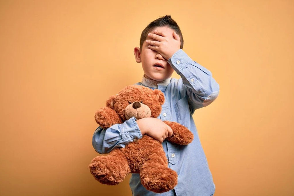  Young boy in a blue shirt covering his eyes while hugging a brown teddy bear against a plain beige background.