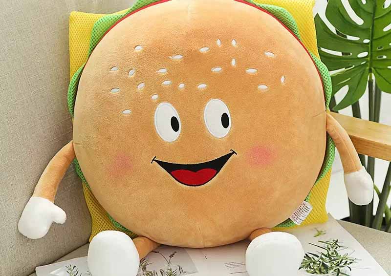 Smiling hamburger-shaped plush toy with arms, legs, and cartoon face sitting on a chair.