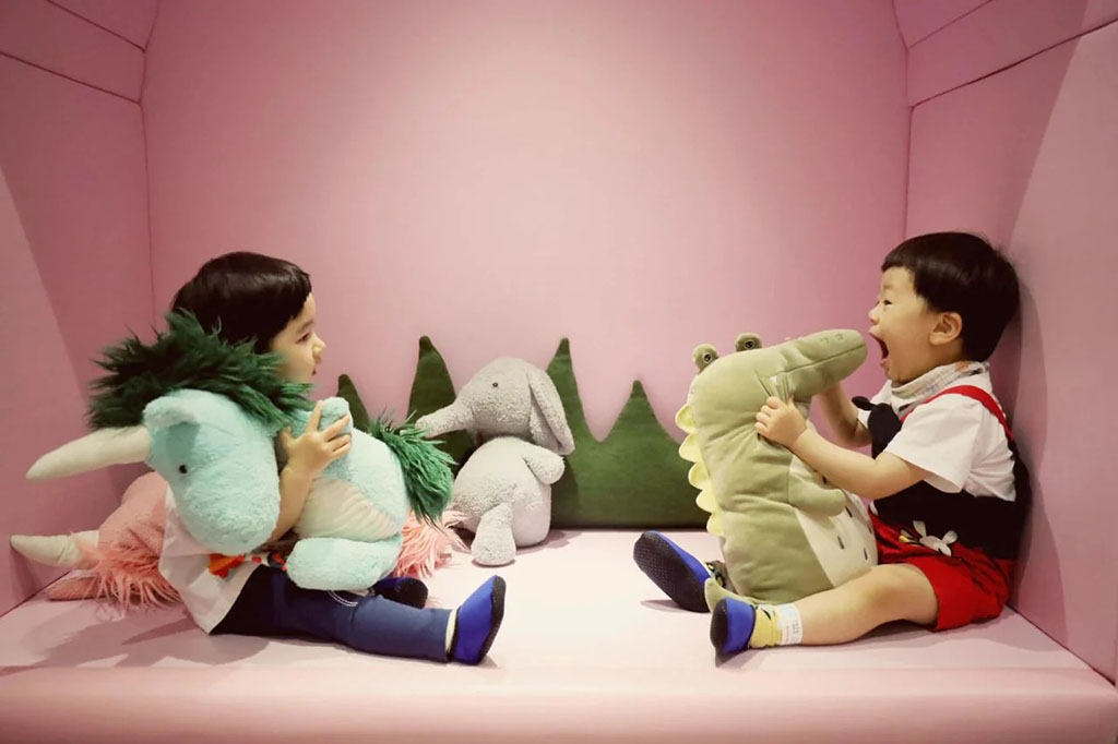 Two young children sitting in a pink play area laughing and holding large dinosaur and unicorn plush toys with other stuffed animals in the background.