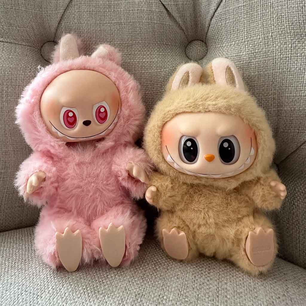  Two collectible Labubu plush dolls in pink and beige furry costumes with large expressive faces and bunny ears sitting on a gray sofa.