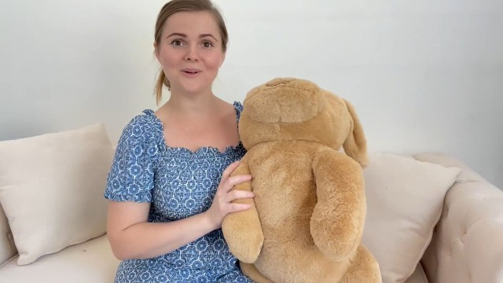 A woman sitting on a sofa holding a large beige plush toy, showing its size and softness for demonstration purposes.