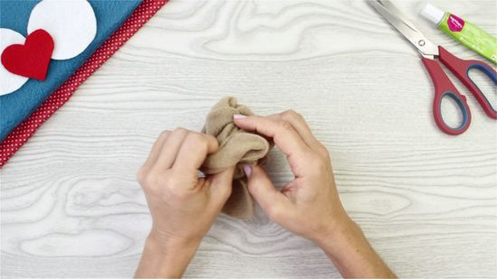 Two hands gathering beige fabric while crafting on a table with scissors and felt pieces nearby.