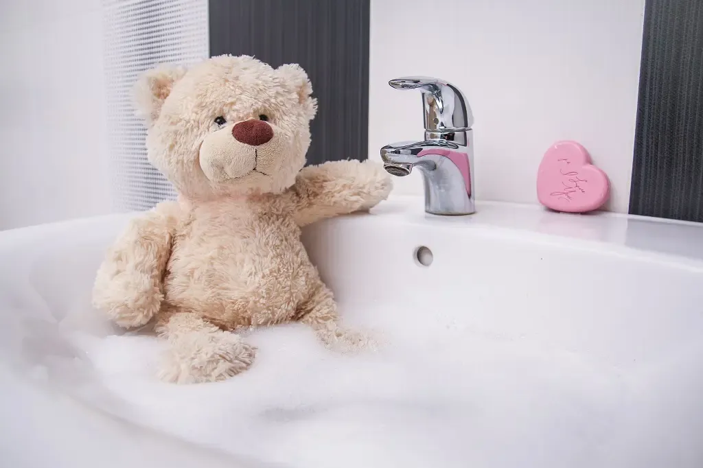 A beige plush teddy bear sitting in a bathroom sink filled with bubbles, leaning against the faucet with a pink heart-shaped soap nearby.