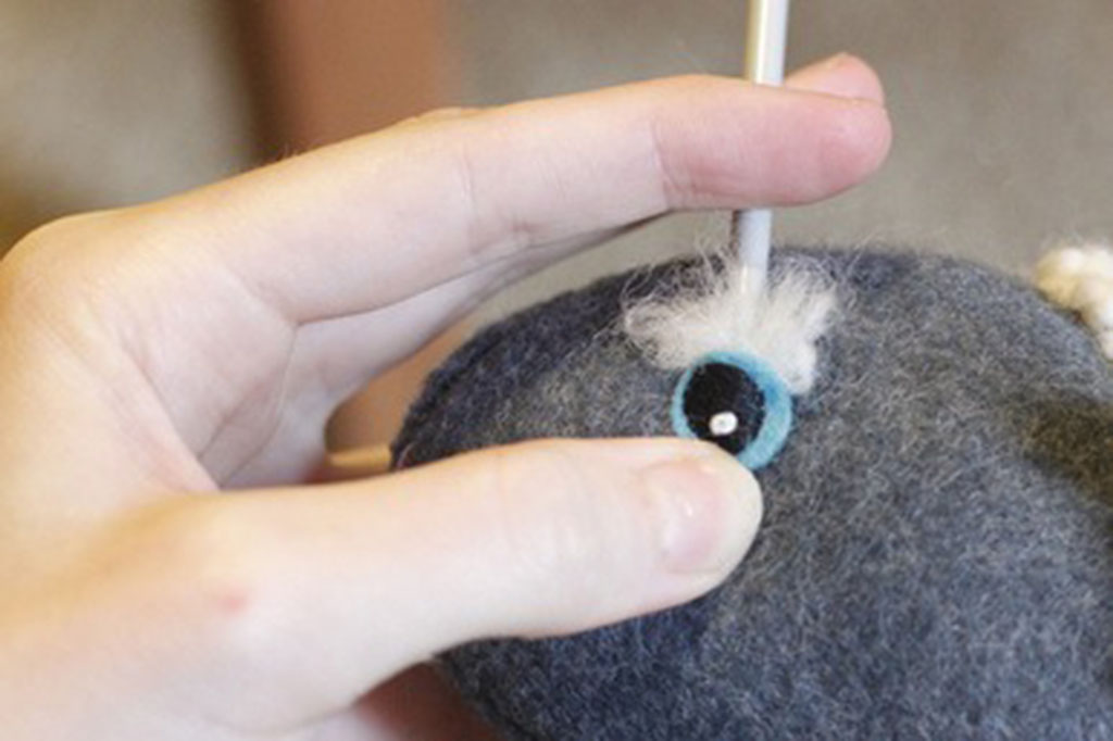 A hand uses a felting needle to attach a wool-felted eye onto a plush toy, showing the needle felting technique used to secure facial features in handmade stuffed animals.