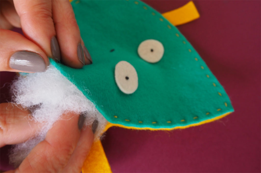 A close-up of hands inserting white fiber stuffing into a small green felt toy with stitched edges and simple felt eyes on a purple background.