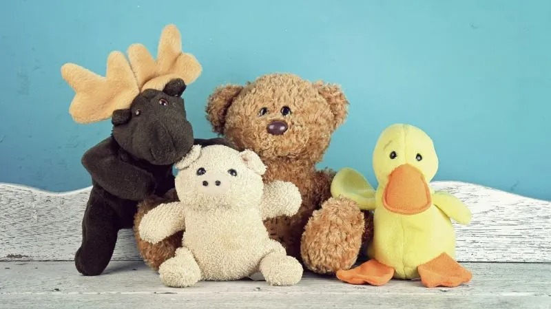 A group of four plush toys—a moose, a brown teddy bear, a white bear, and a yellow duck—sitting together against a blue background on a wooden surface.