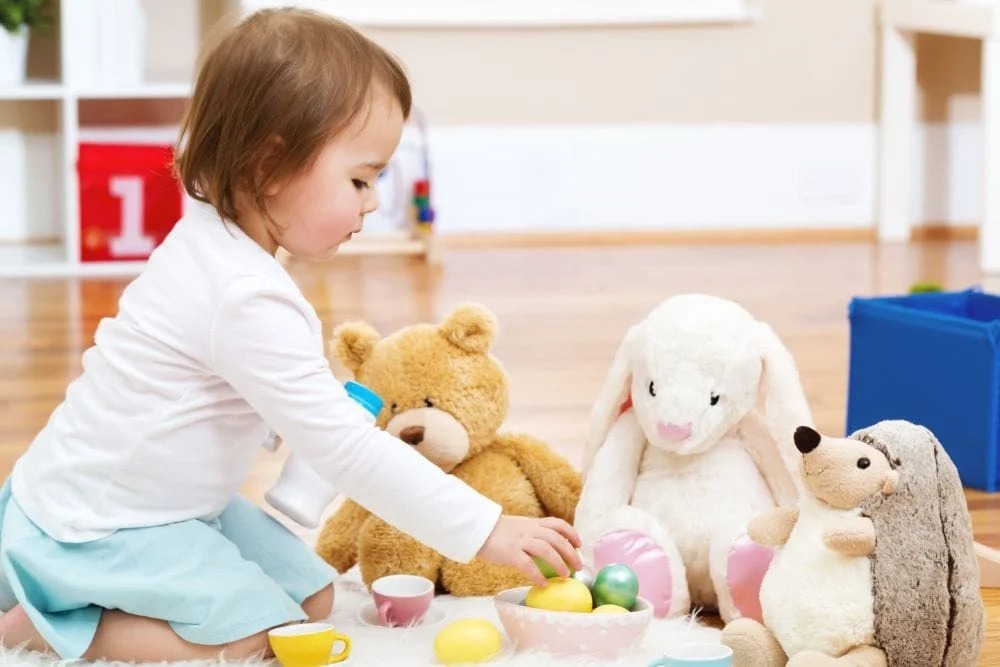 Young child sitting on the floor hosting a pretend tea party with a teddy bear, white bunny plush, and hedgehog plush toys arranged in front.
