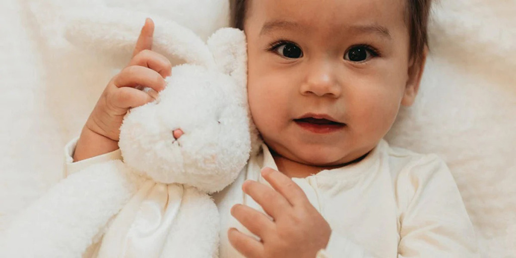 A baby lying on a soft surface while gently holding a white plush bunny, looking toward the camera with a calm expression.