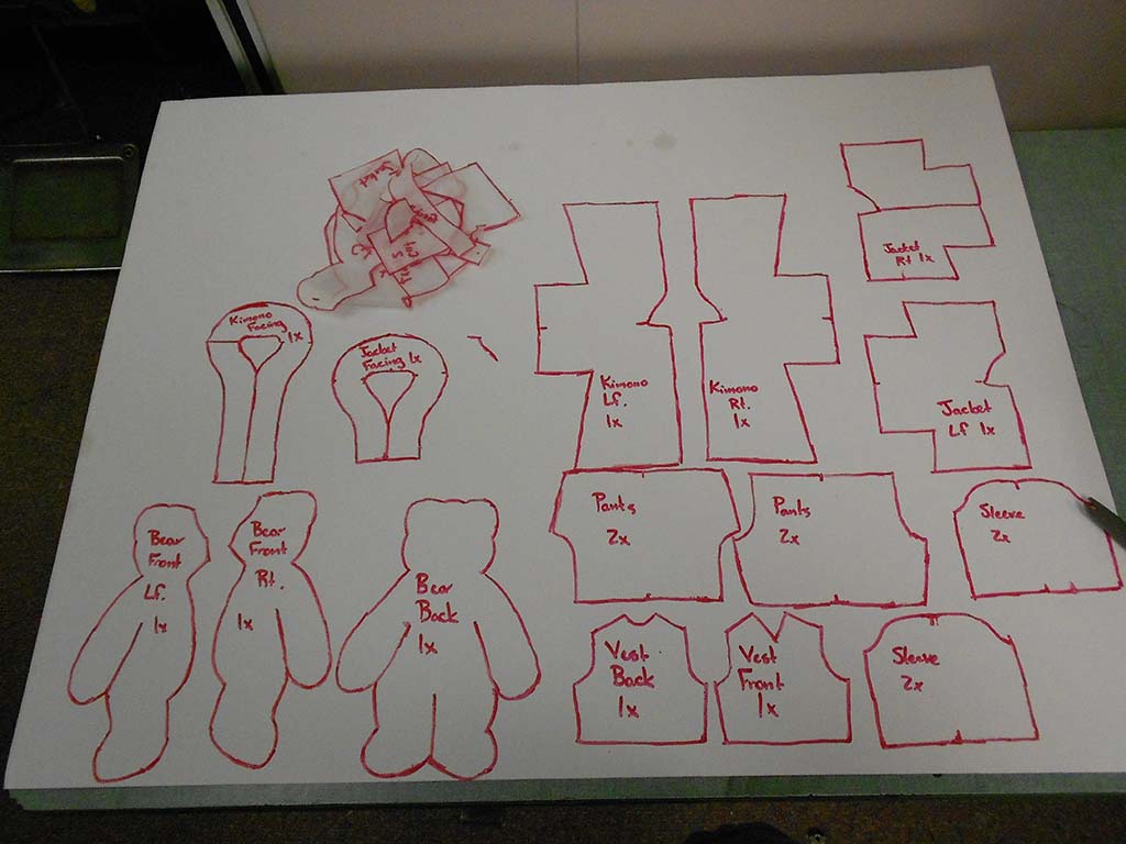 Large white sheet covered with red-outlined pattern pieces for sewing a plush bear and its clothing, including body fronts, back, sleeves, pants, vest, kimono pieces, and pocket facings.