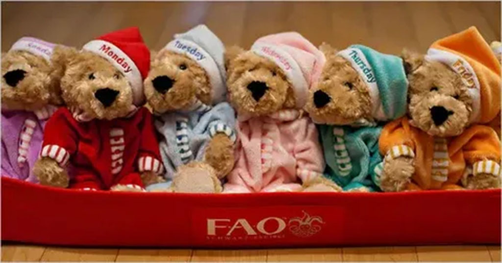 A row of FAO Schwarz teddy bears dressed in colorful day-of-the-week outfits, sitting together in a red display tray.