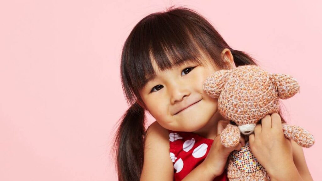 A young child smiling and hugging a knitted teddy bear against a soft pink background.