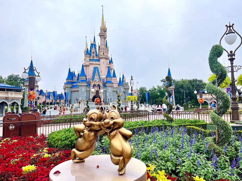 Golden Chip ’n’ Dale statue in front of a colorful flower garden with the iconic Disney castle in the background at a theme park.